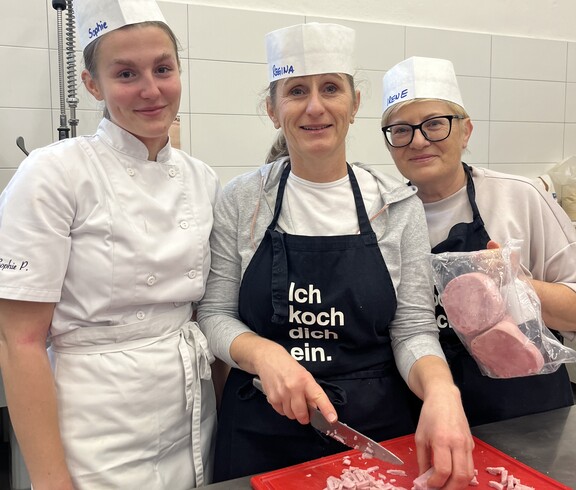 Drei Frauen schauen in die Kamera. Eine von ihnen schneidet gerade Schinken in Würfel. 