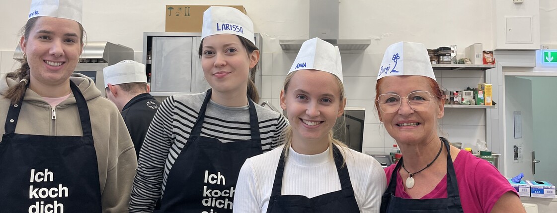 4 junge Frauen in Kochmützen und Schürzen mit dem Aufdruck "ich koch dich ein" posieren in einer Küche für die Kamera. 