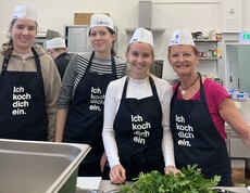 4 junge Frauen in Kochmützen und Schürzen mit dem Aufdruck "ich koch dich ein" posieren in einer Küche für die Kamera. 