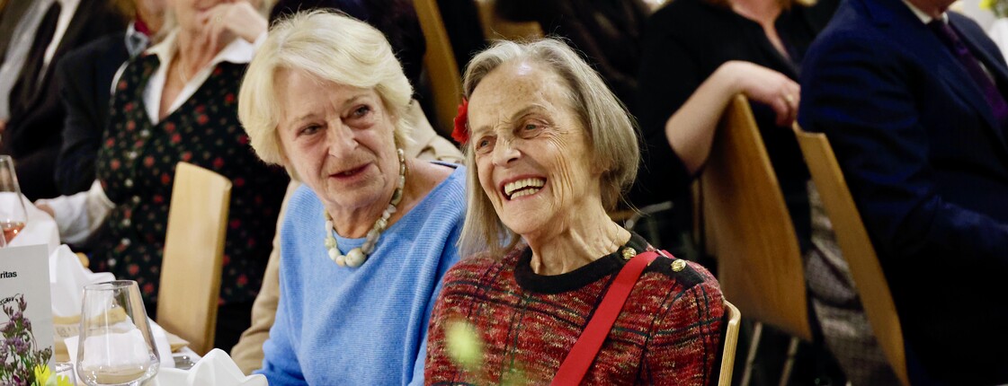 Mehrere Personen sitzen an gedeckten Tischen. Im Fokus ist eine Dame, die lächelt. 